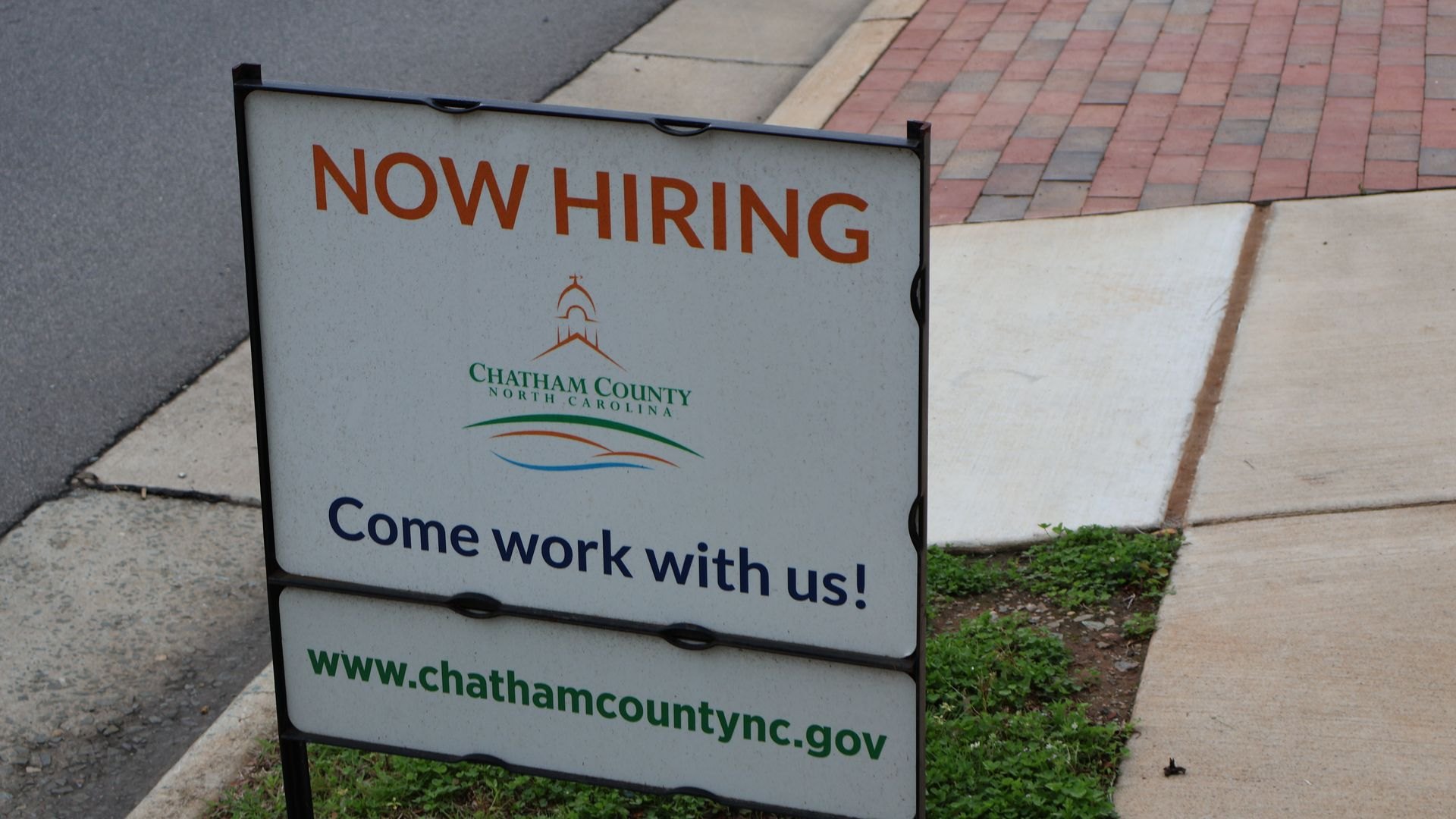 A "Now Hiring" sign on a sidewalk